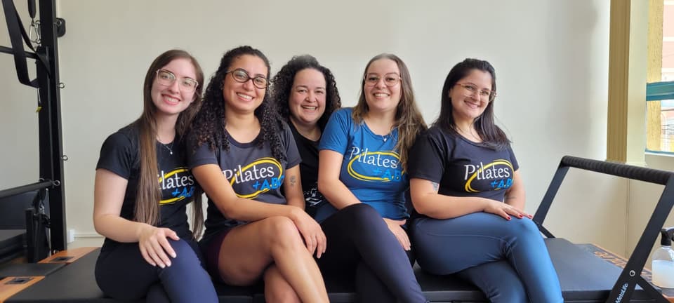 Cinco instrutoras do Pilates+ABC sorrindo, sentadas lado a lado em um aparelho do estúdio.
